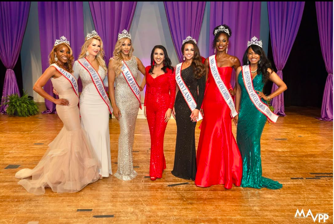 Seven women in evening gowns and sashes stand on stage with purple curtains in background.