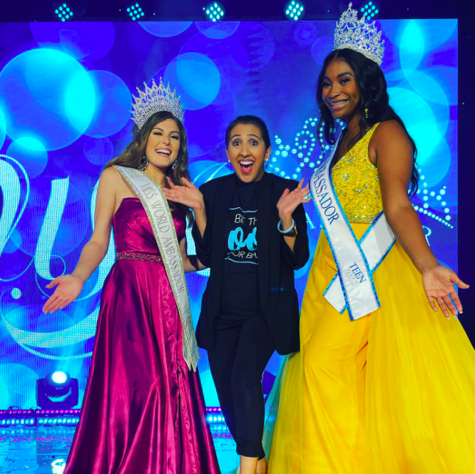 Three women on stage, two wearing crowns and sashes, one in a black outfit, all smiling with a blue background.