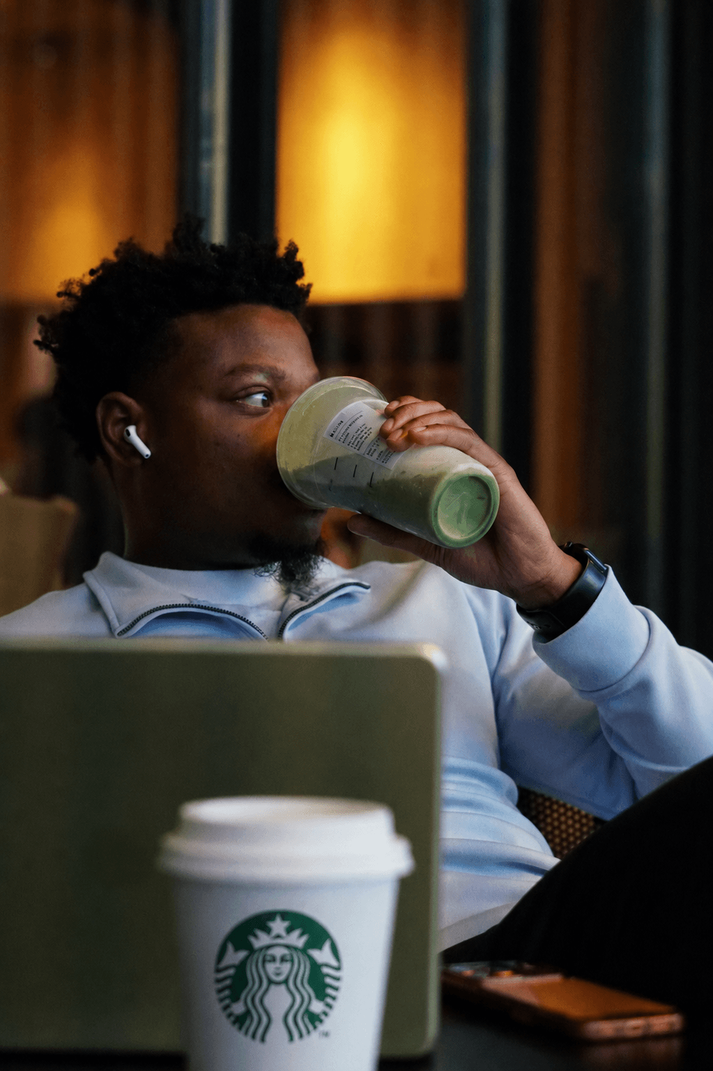 Person with short curly hair drinking a green smoothie in a cafe, with a Starbucks cup and laptop nearby.