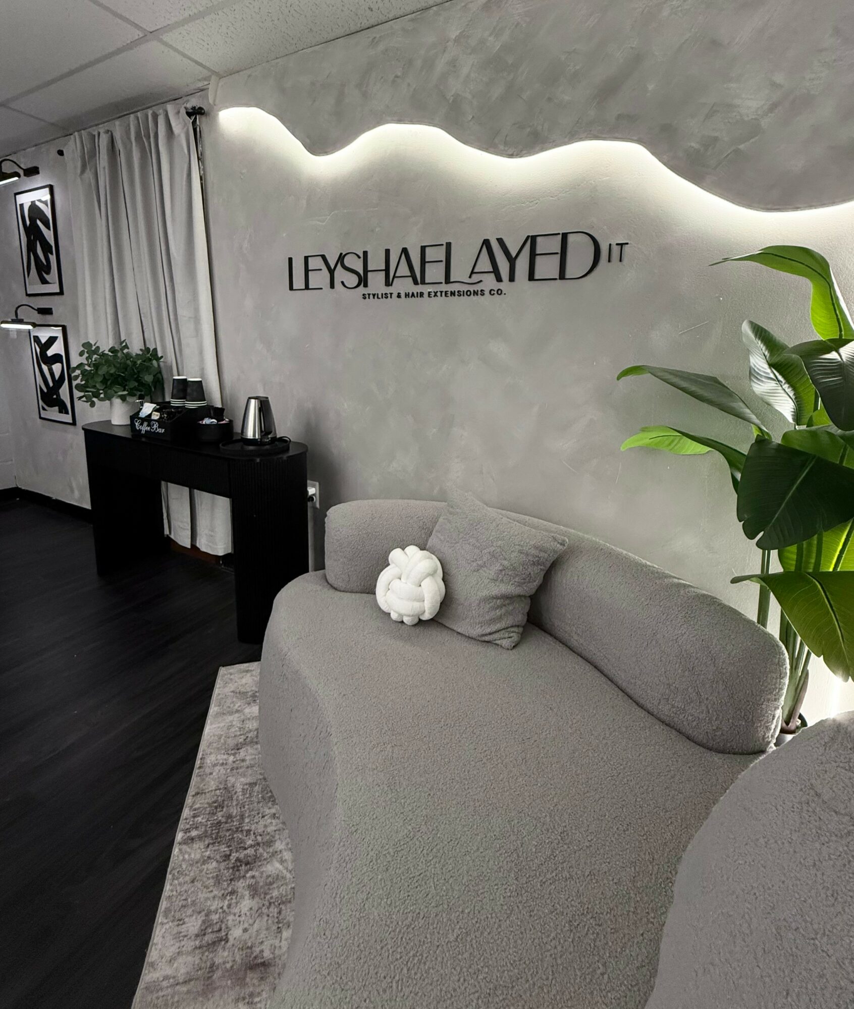 Reception area with a gray sofa, a large plant, and a wall with the sign 'LESHAYLAAYED' illuminated above.