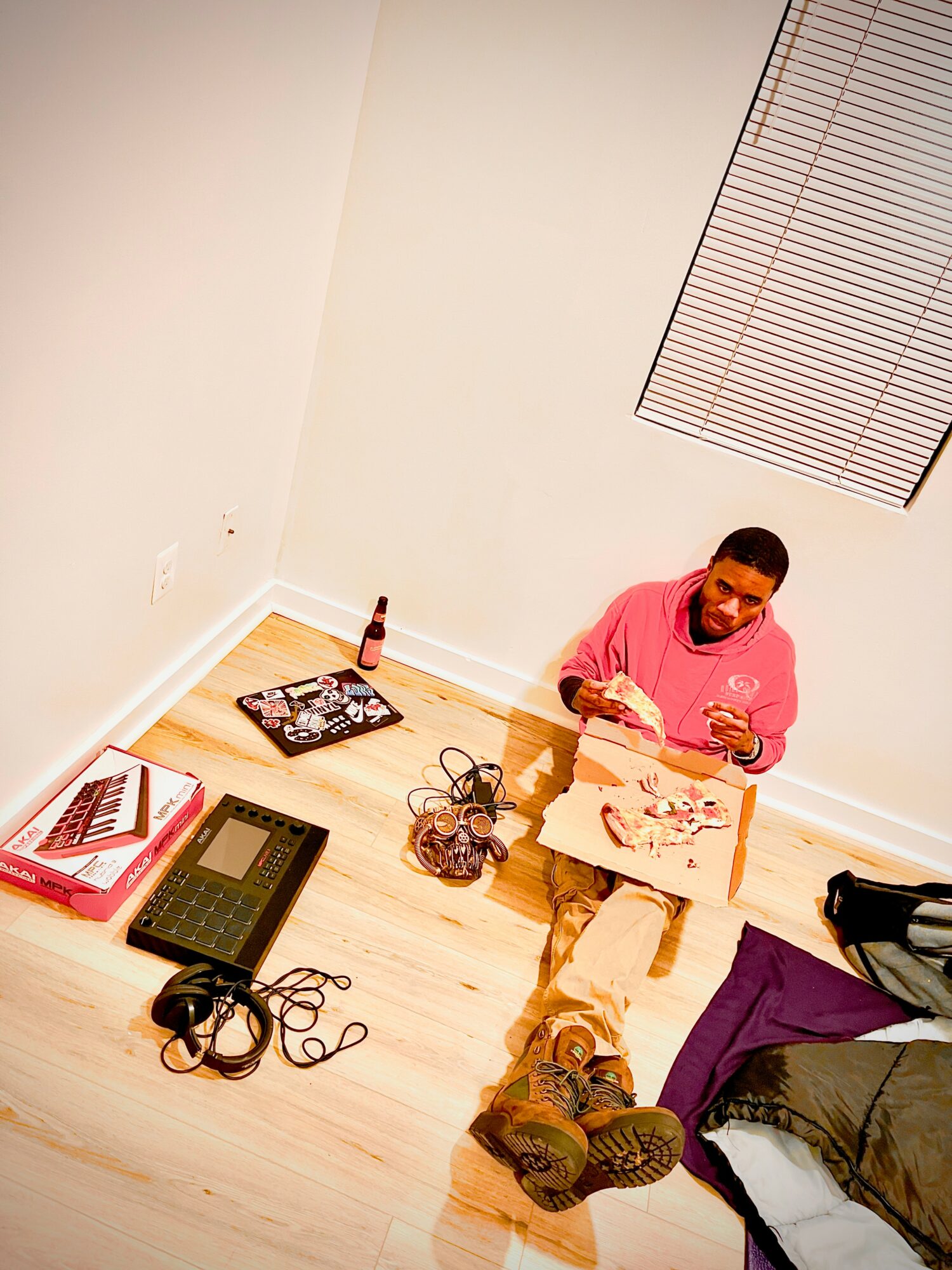 Person sitting on the floor with a pink hoodie, surrounded by books, a bottle, and electronic devices, in a room with light-colored walls.