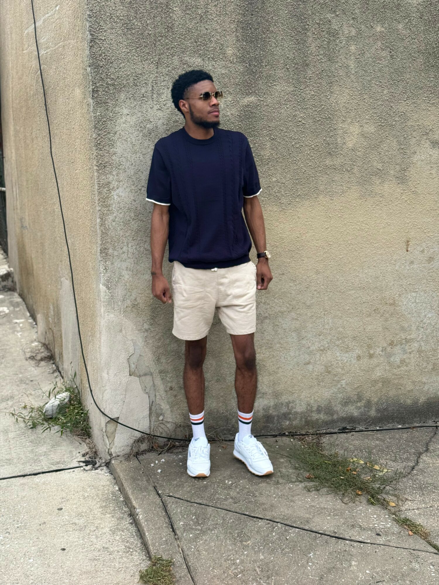 Young man standing against a textured concrete wall, wearing a navy T-shirt, beige shorts, white sneakers, and striped socks.