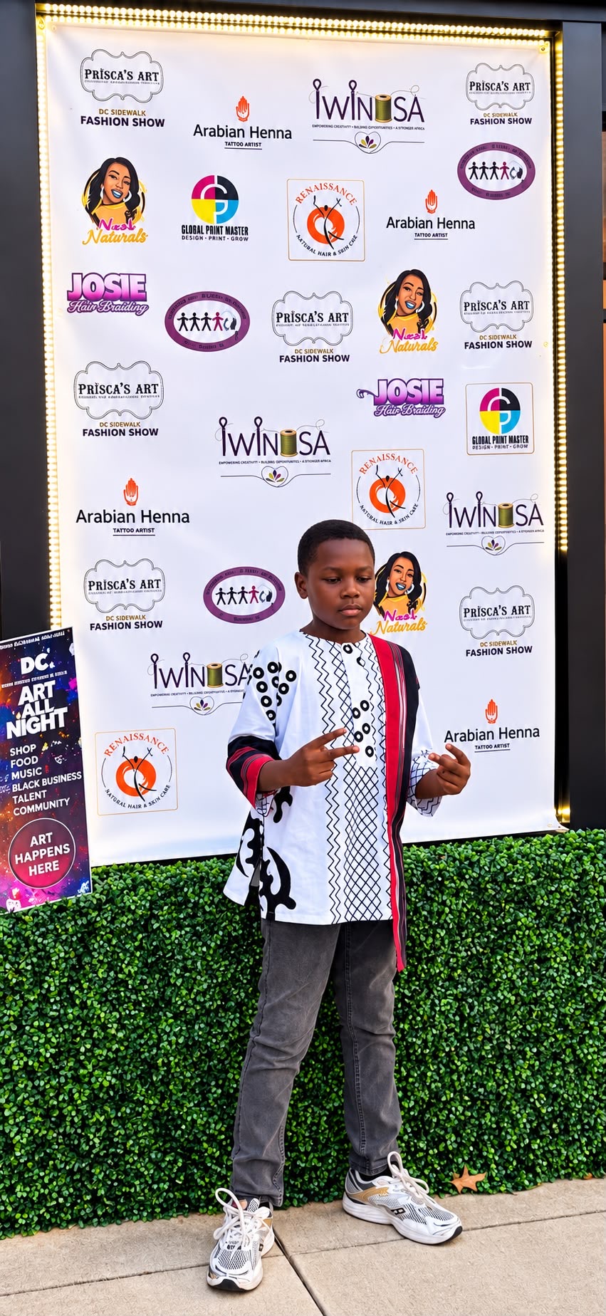 Young boy standing outdoors in front of a large event banner, holding a phone, wearing a white and black jacket, gray pants, and sneakers.