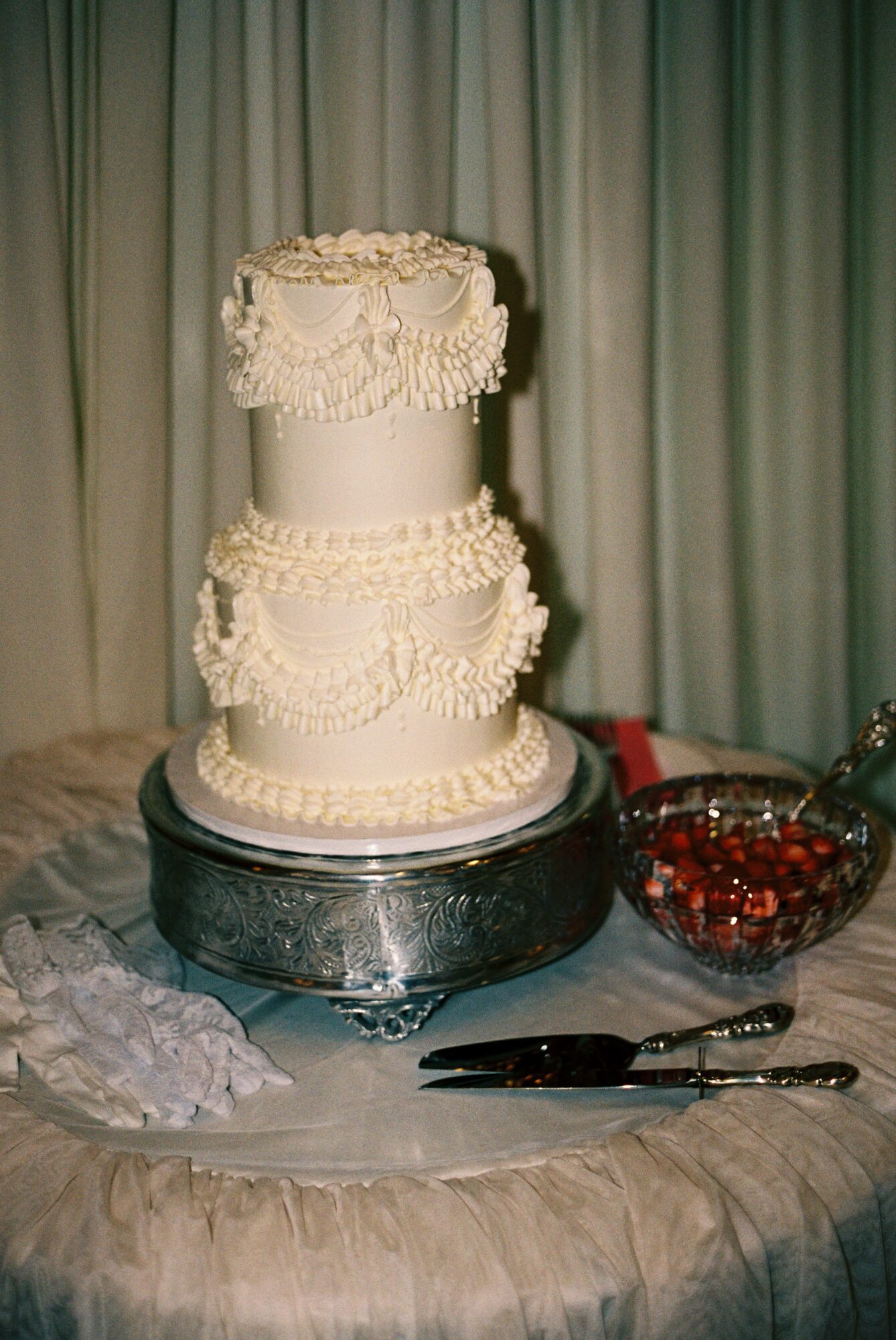 Three-tiered wedding cake with decorative icing, on a silver stand, with a bowl of cherries and a knife nearby, against curtains.
