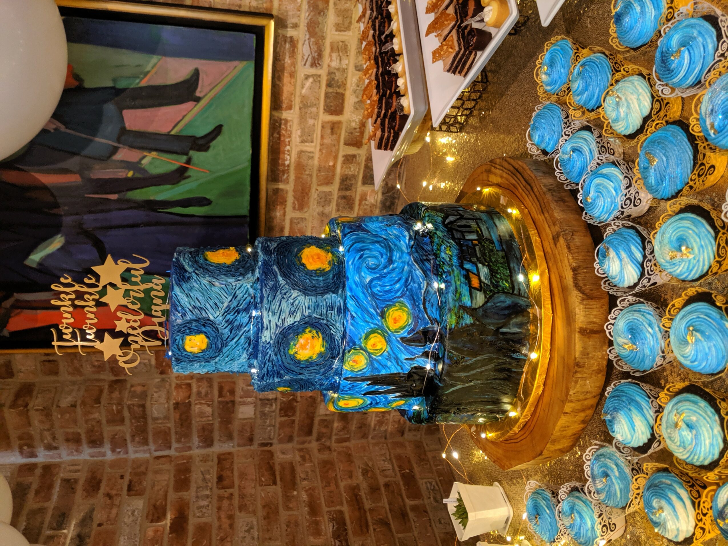 Blue cake decorated with yellow flowers and a sunflower, surrounded by blue and white cupcakes, on a gold cake stand.
