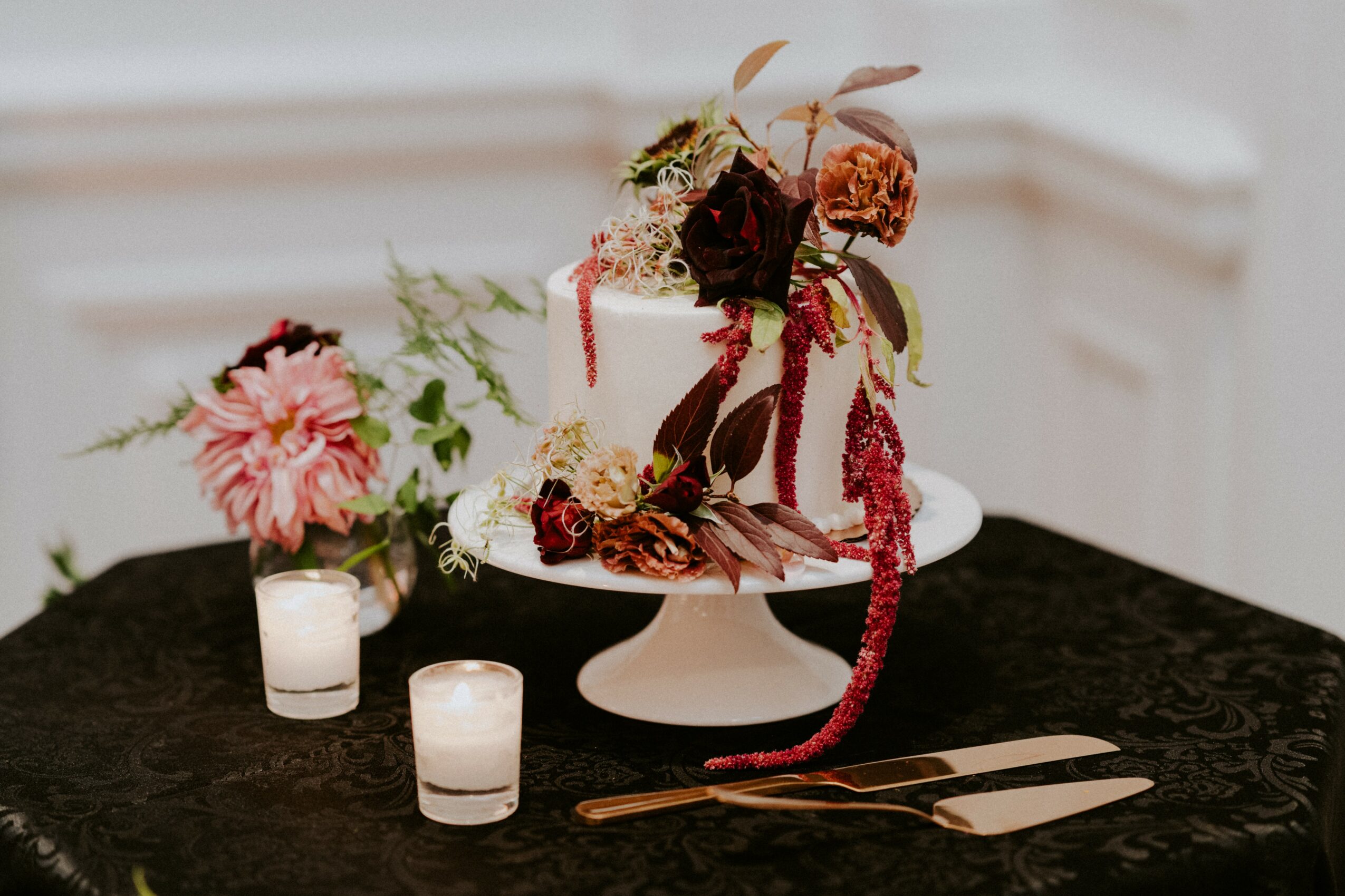 Decorative cake with flowers and foliage on a table, surrounded by candles and utensils, in a bright room.