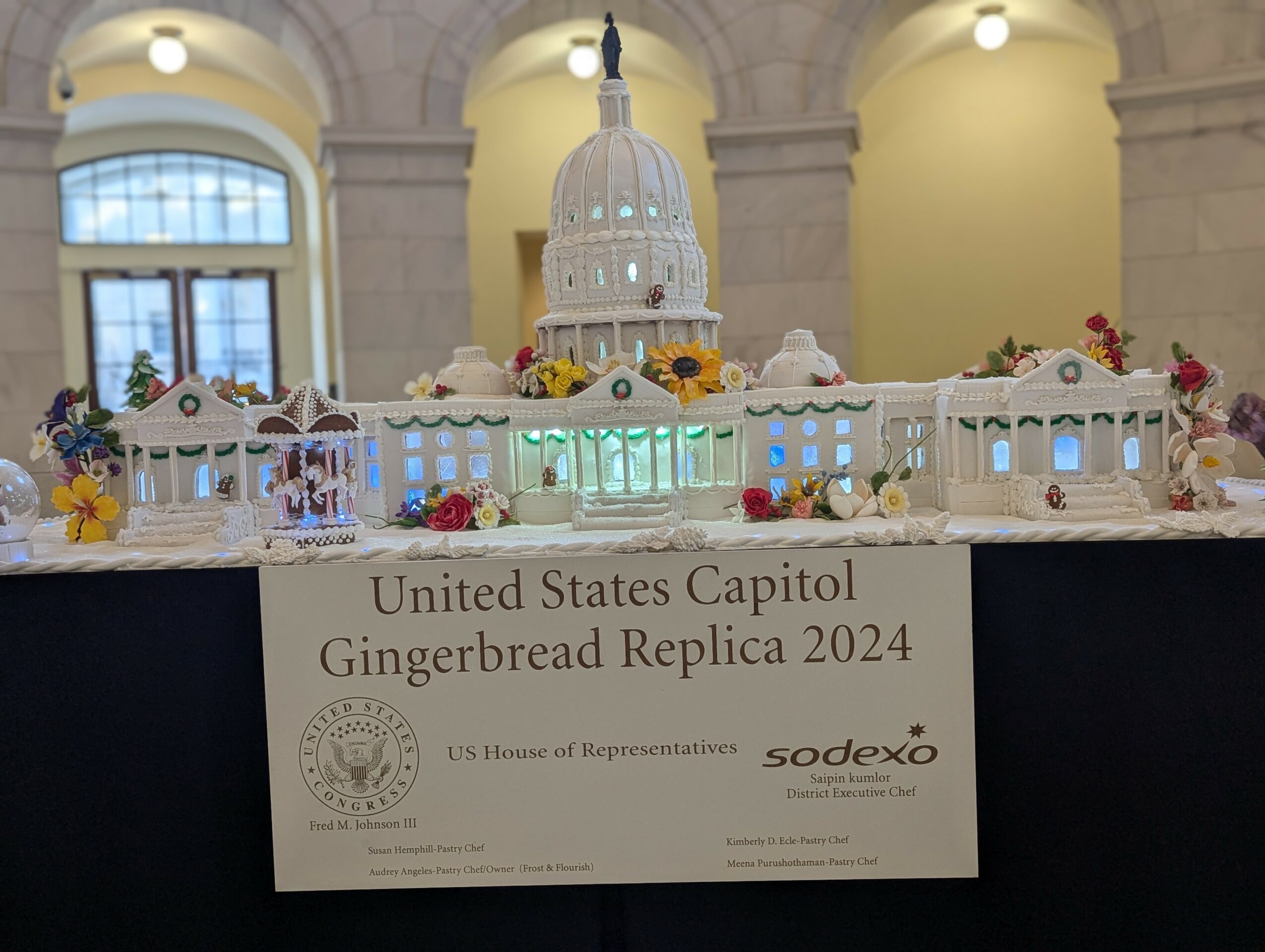 Gingerbread replica of the United States Capitol building with flowers, displayed on a table inside a building with arched windows.
