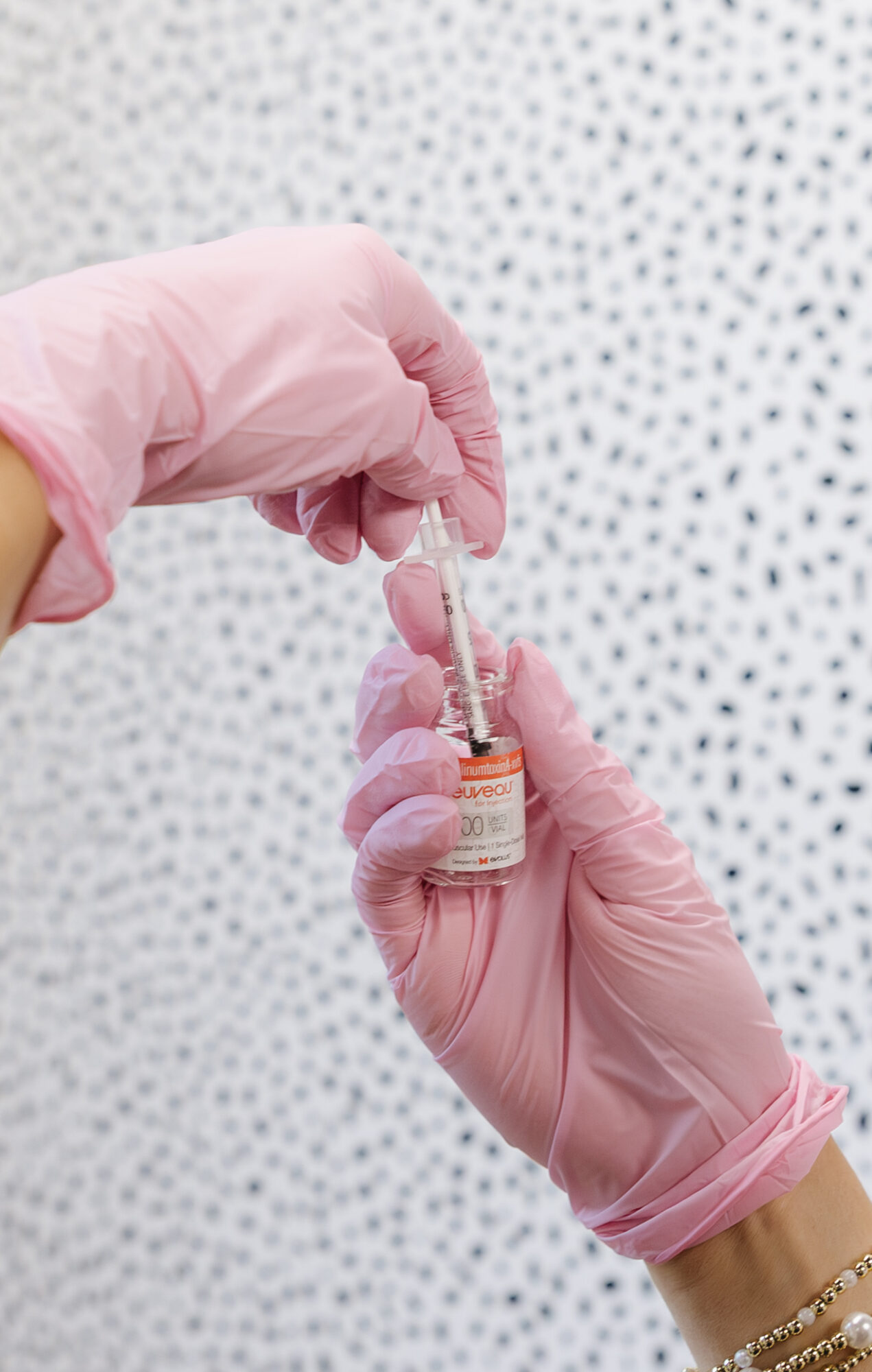Person wearing pink gloves holding a small vial with a syringe, against a dotted background.