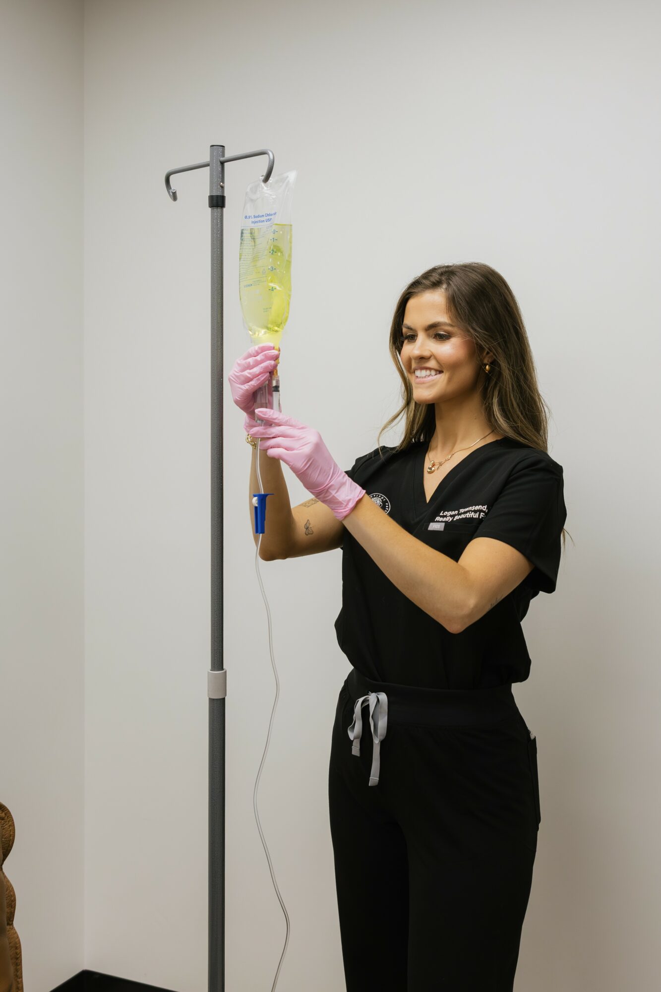 Young woman in black scrubs and pink gloves prepares an IV bag in a clinical setting.