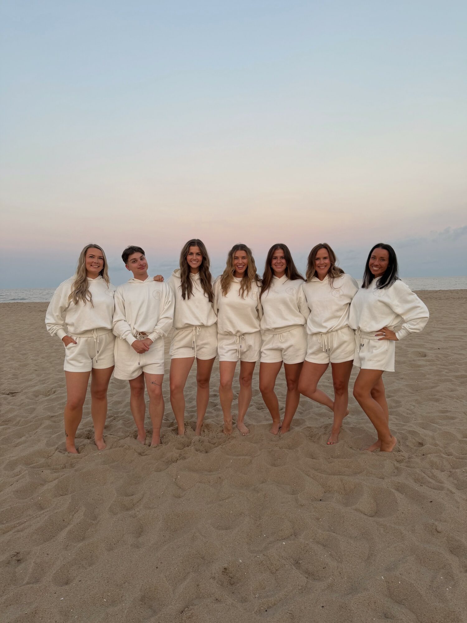 Seven women stand on a beach, wearing matching white outfits, with the ocean and sky in the background.