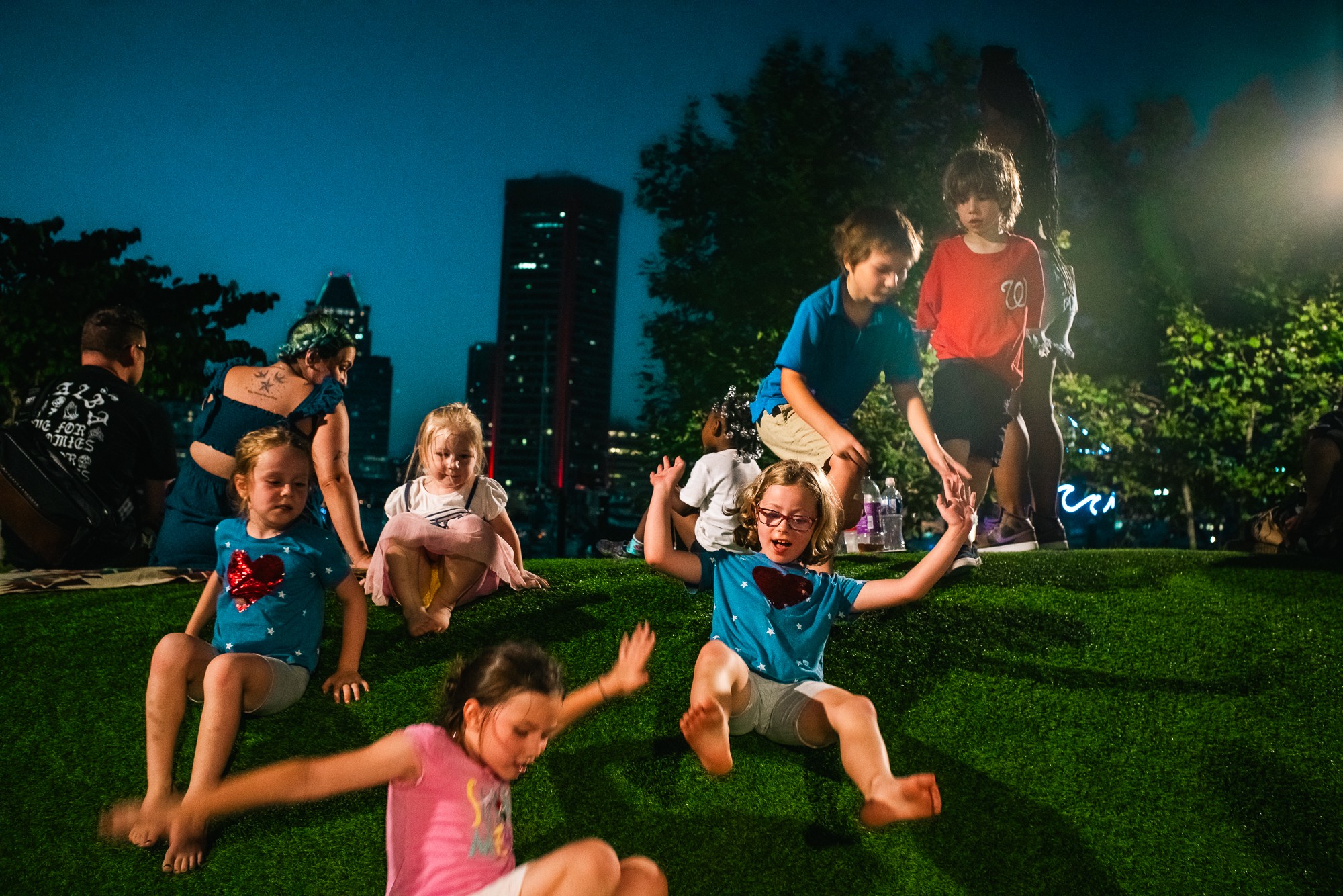 Children playing on grassy steps outdoors at night with city buildings and trees in background.