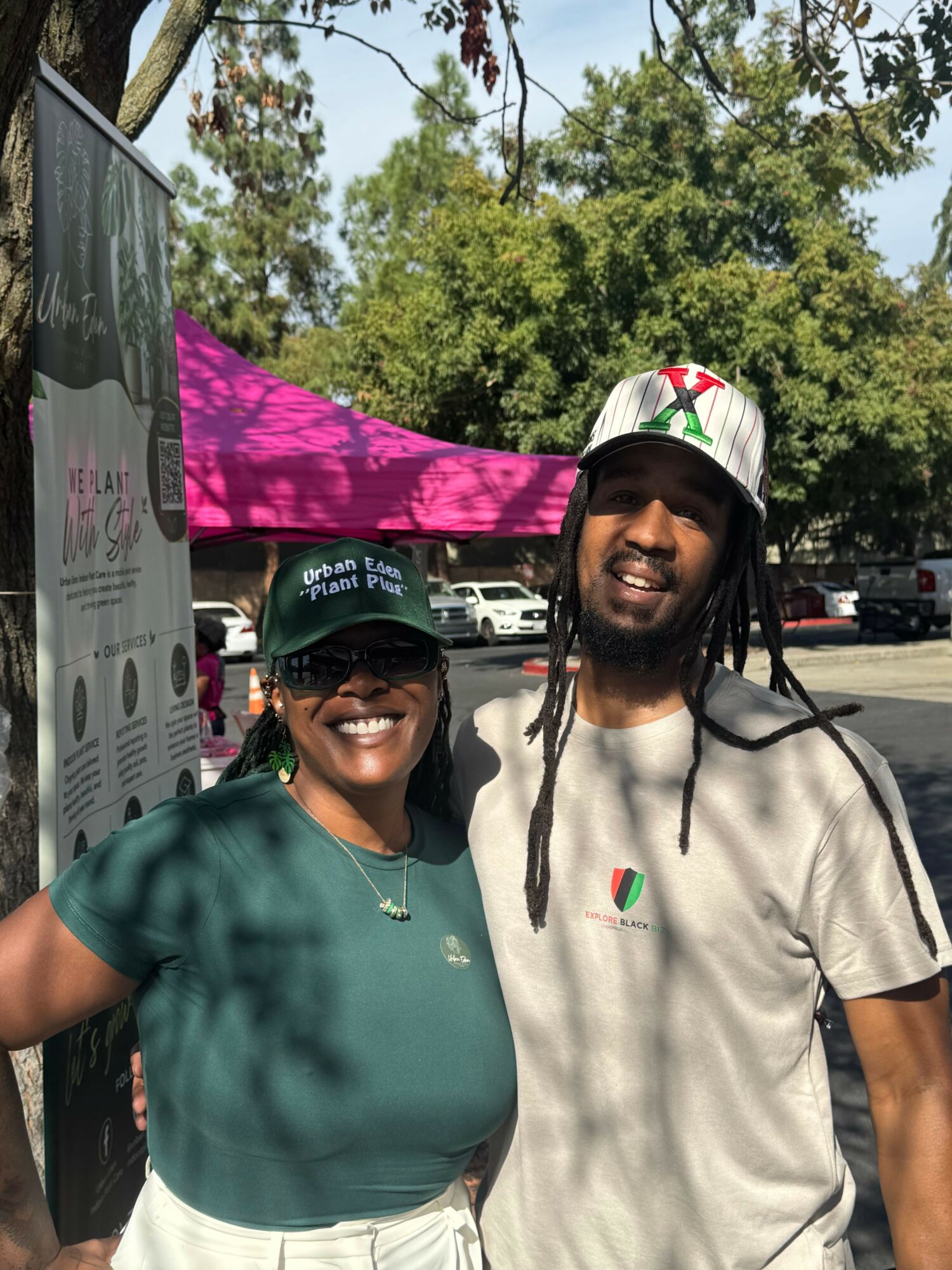 Two women smiling outdoors, one wearing a green cap and the other with a hat, standing near a pink umbrella.