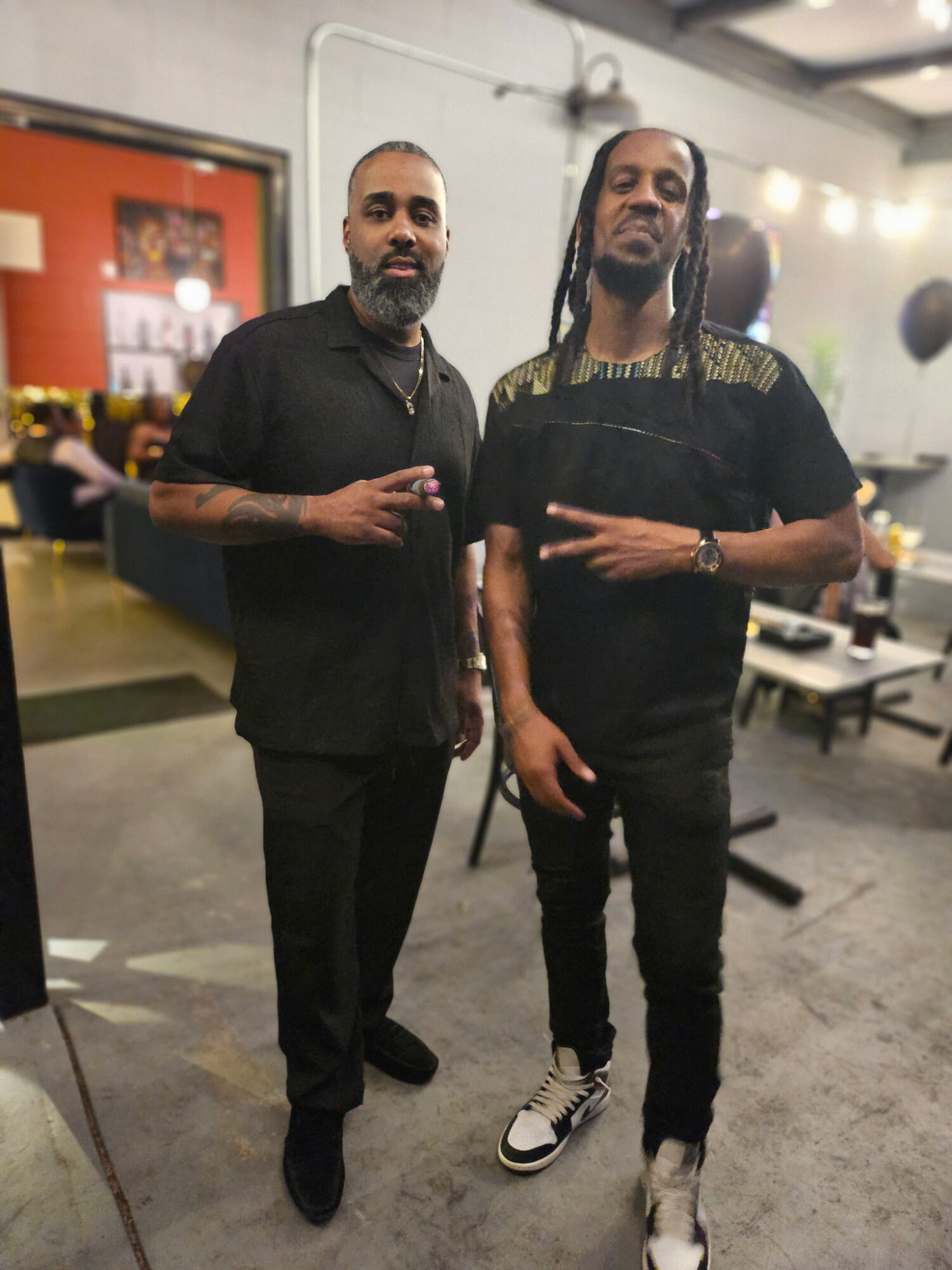 Two men standing indoors, one with a beard and tattoos, the other with long braids, both wearing black clothing.