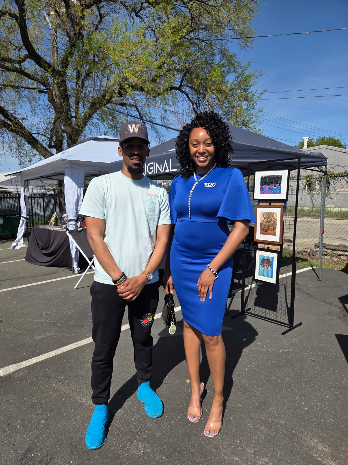 A man and woman stand outdoors in front of an art display, smiling, with trees and a tent in the background.