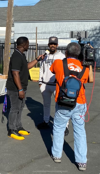 Three men are standing outdoors, one with a camera, in a parking lot with a fence and building in background.