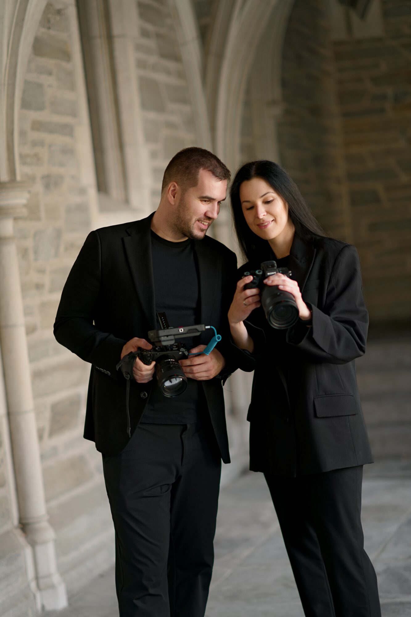 Two people in black suits look at a camera together outside a stone building with arches.