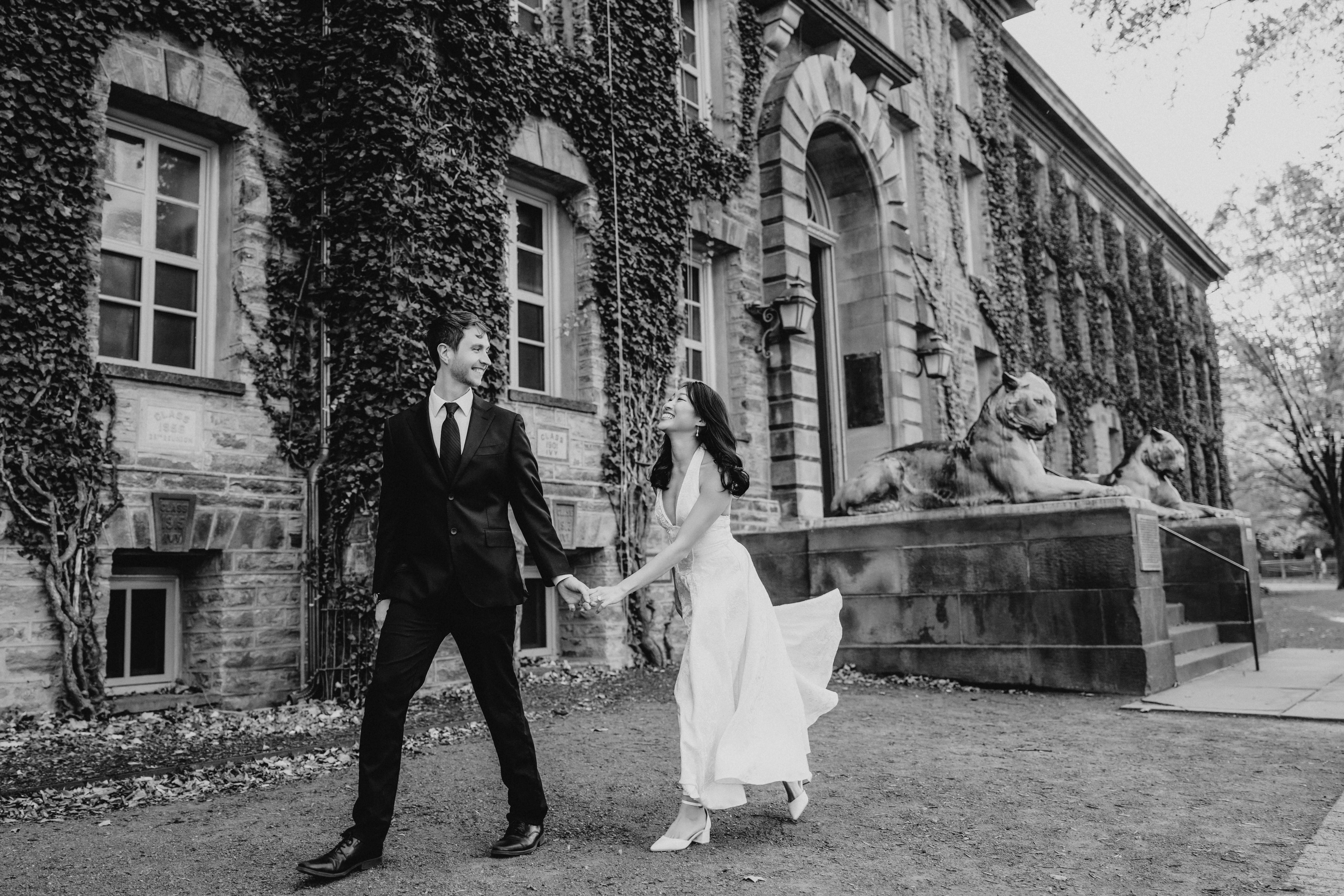 A couple holding hands walking outside a historic building with lion statues, trees, and ivy-covered walls.