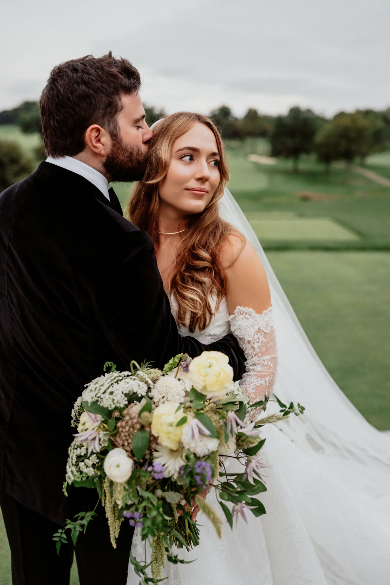 Bride and groom standing outdoors, bride holding a large bouquet, groom in a black suit, green landscape background.