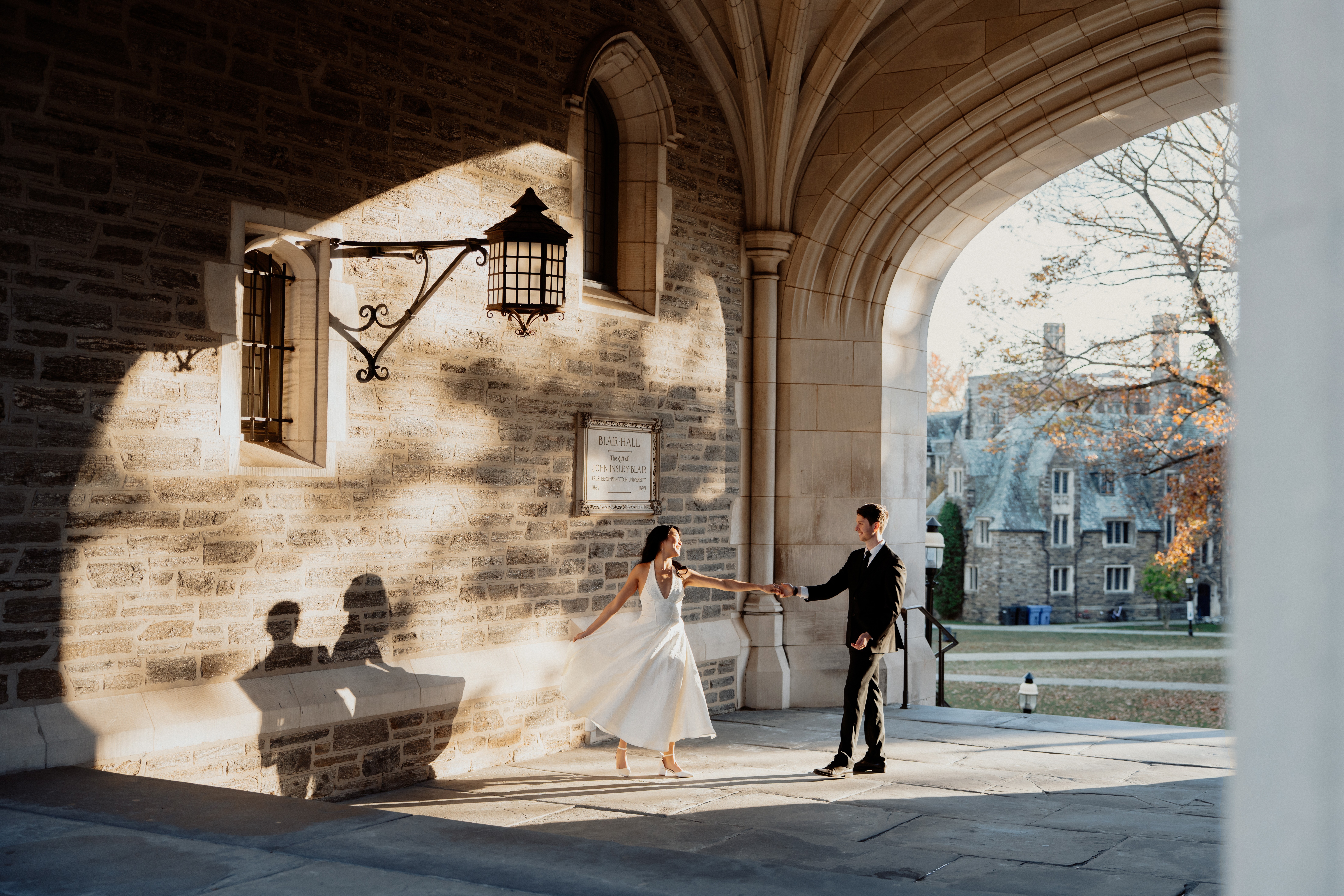 A bride and groom holding hands under an archway with sunlight and shadows, outside a stone building, with trees and buildings in background.