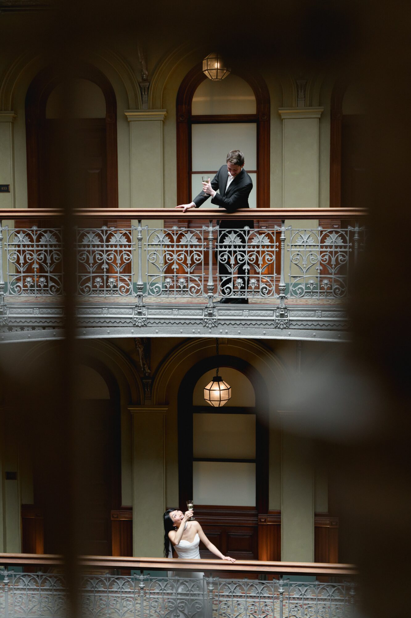 Two people sitting on a balcony inside a building with ornate railing, one on the upper level and one on the lower level.
