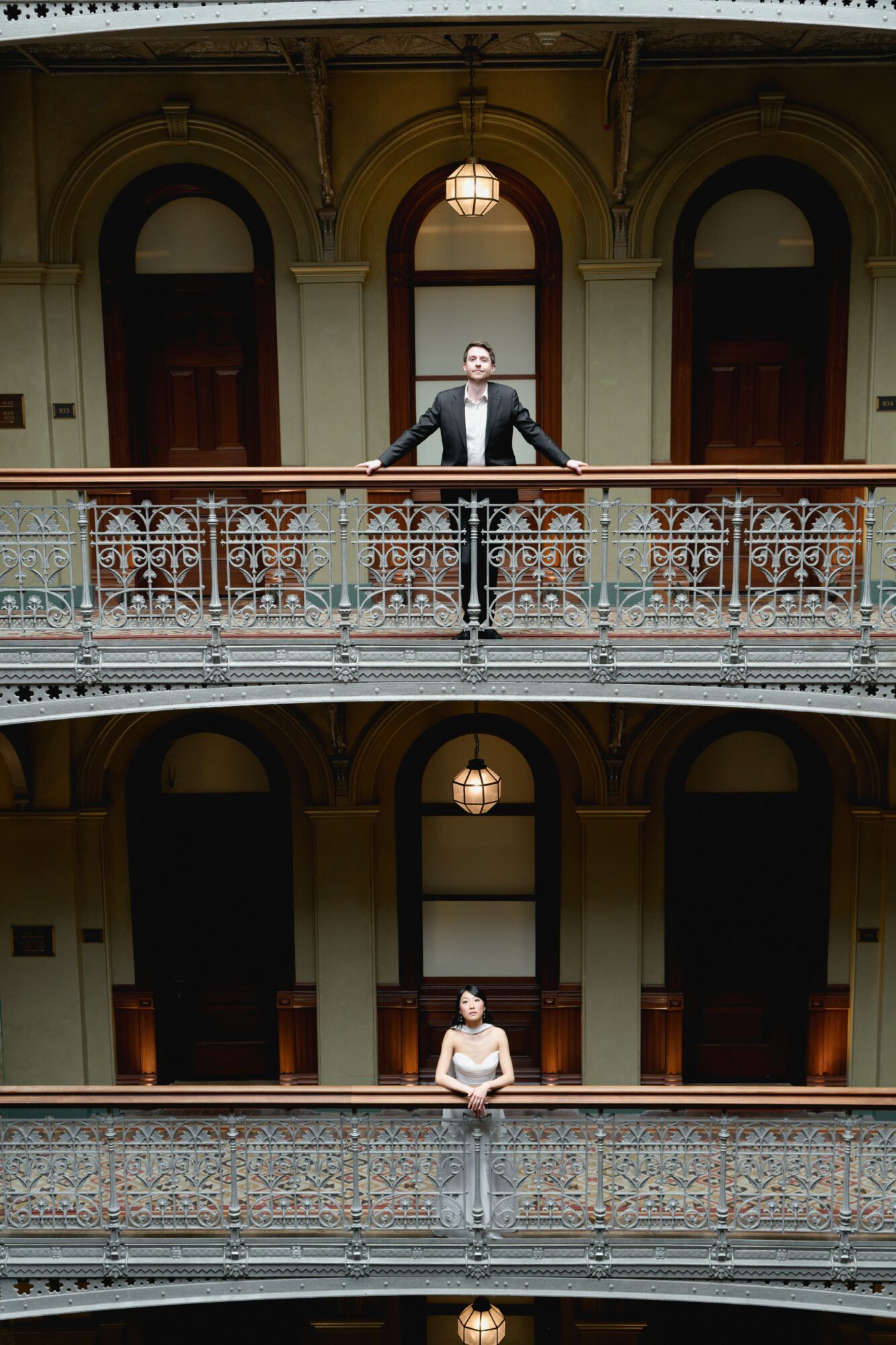 Man in suit standing on balcony railing, woman sitting on lower balcony, ornate interior with arches and hanging lights