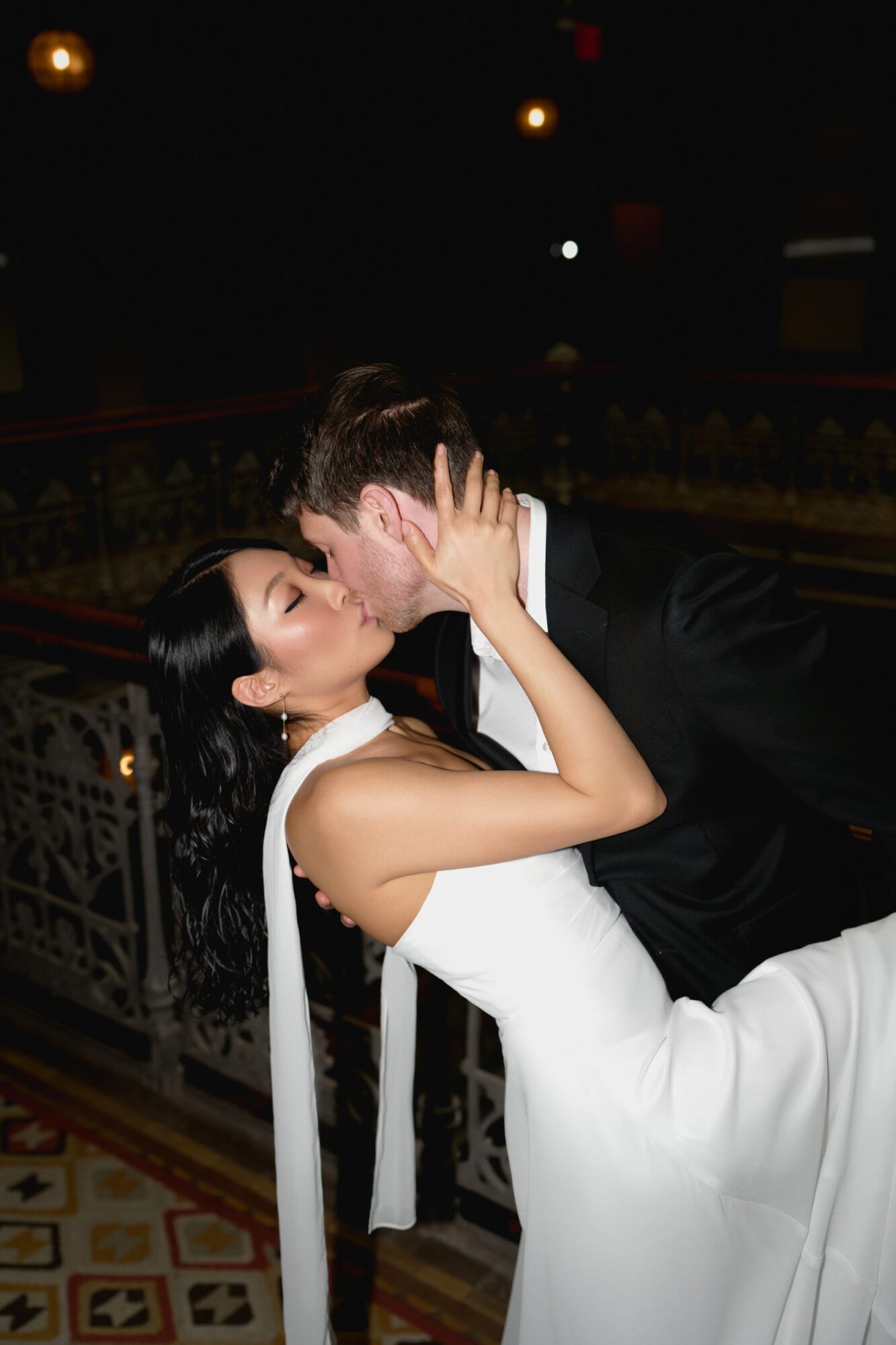 A couple in formal attire sharing a kiss indoors at night, with ornate decor in the background.