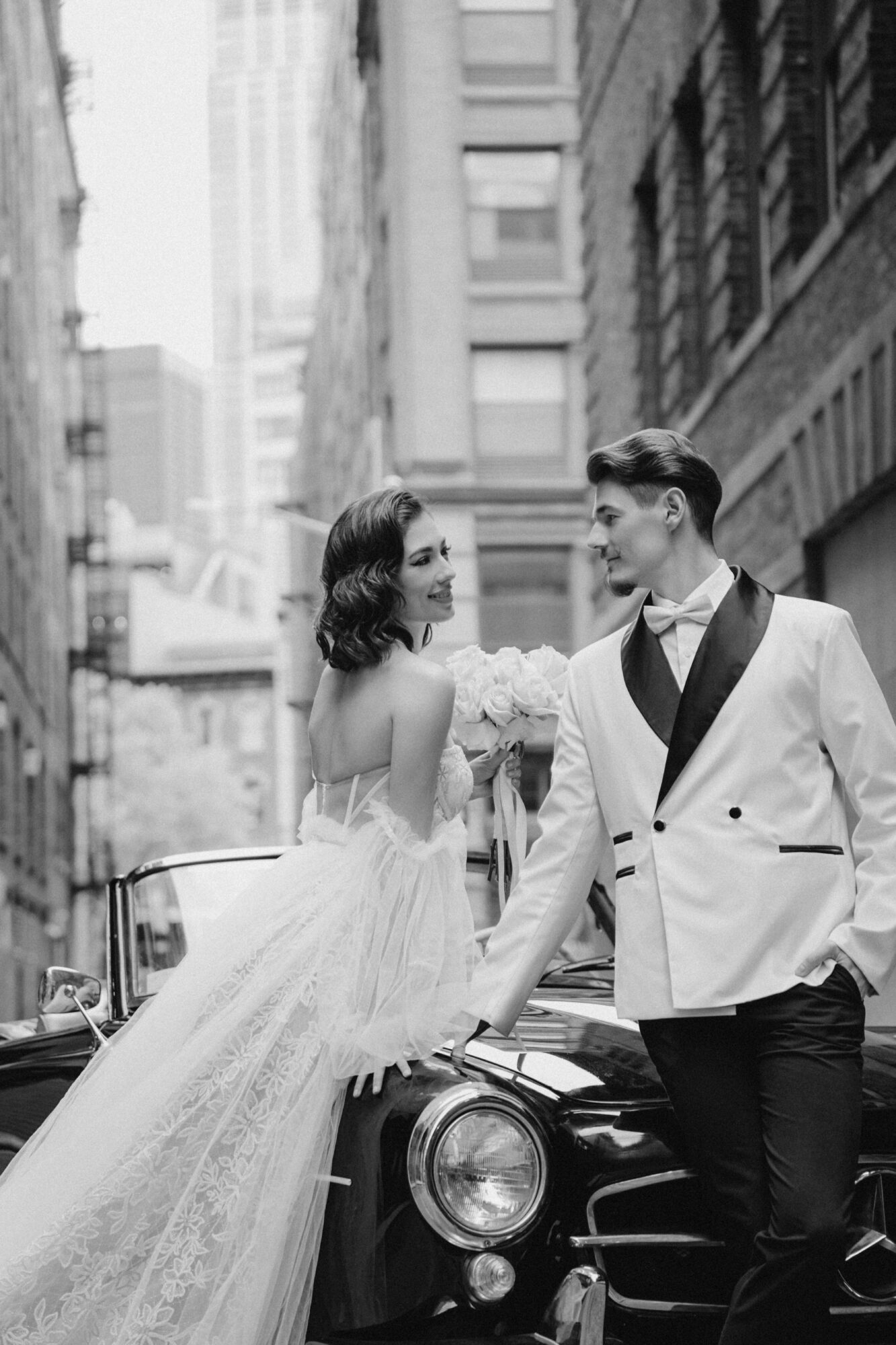 Couple in formal attire leaning on a vintage car in city street, smiling at each other, tall buildings in background.
