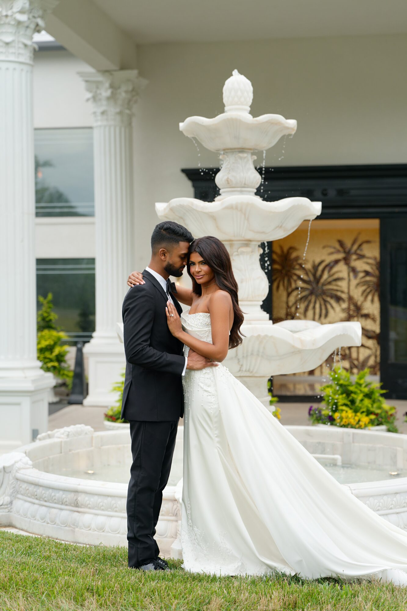 A couple in wedding attire stands close in front of a multi-tiered fountain outdoors, holding hands and looking at each other.