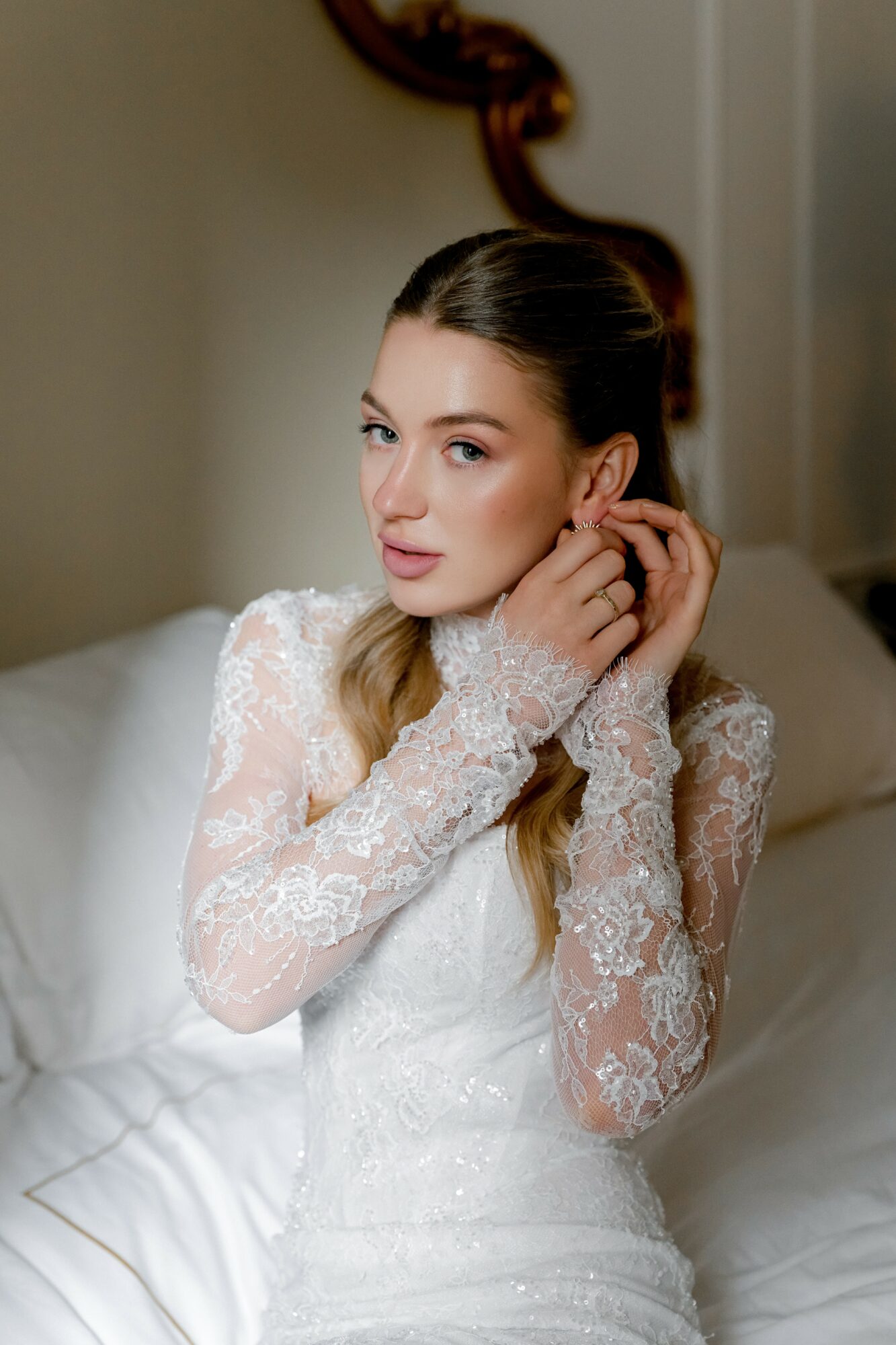 Young woman in lace dress adjusting earring, sitting on bed with white bedding, indoors.