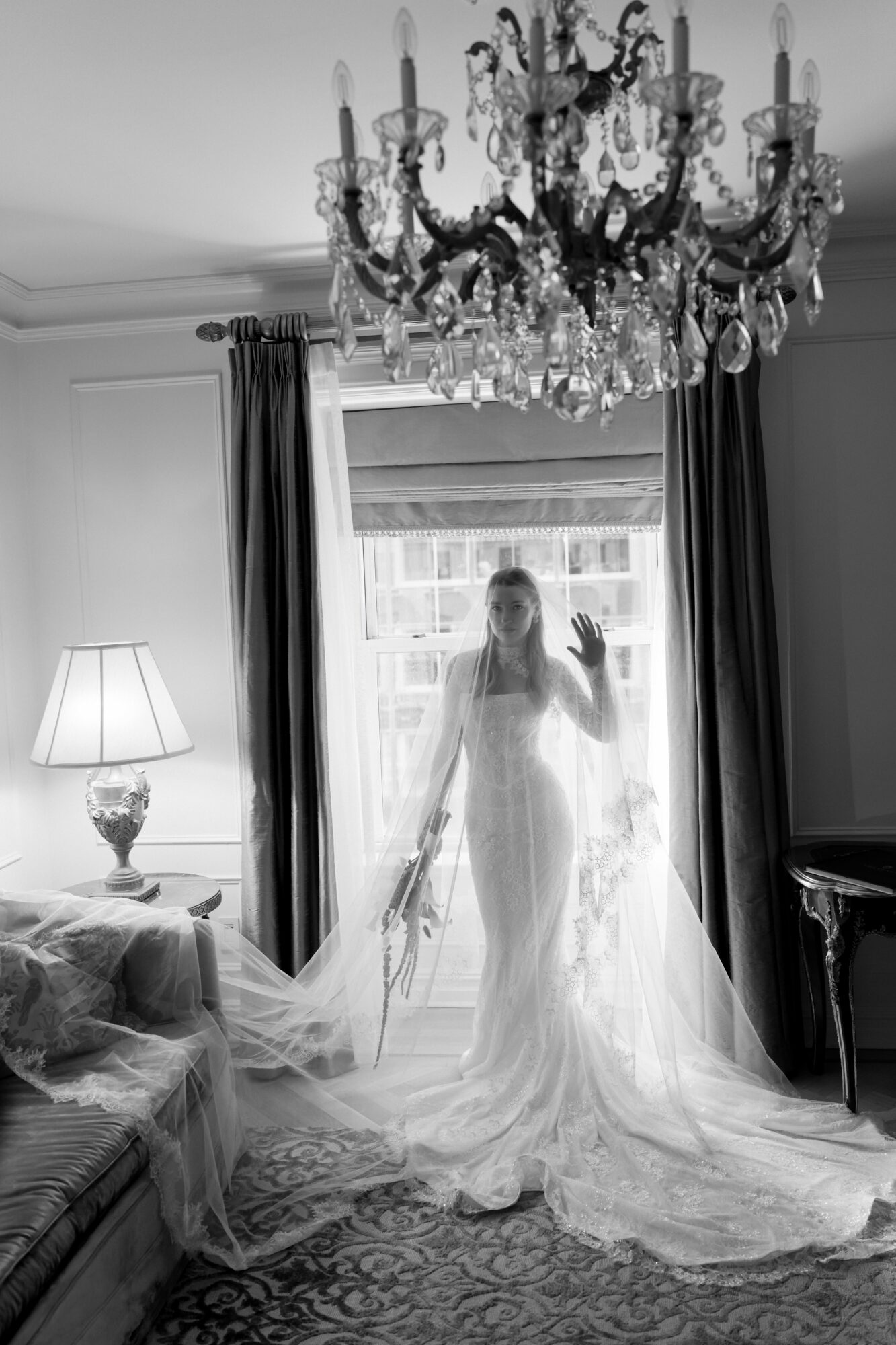 A woman in a wedding dress stands in front of a window with curtains, holding a bouquet, in a room with a chandelier and lamp.