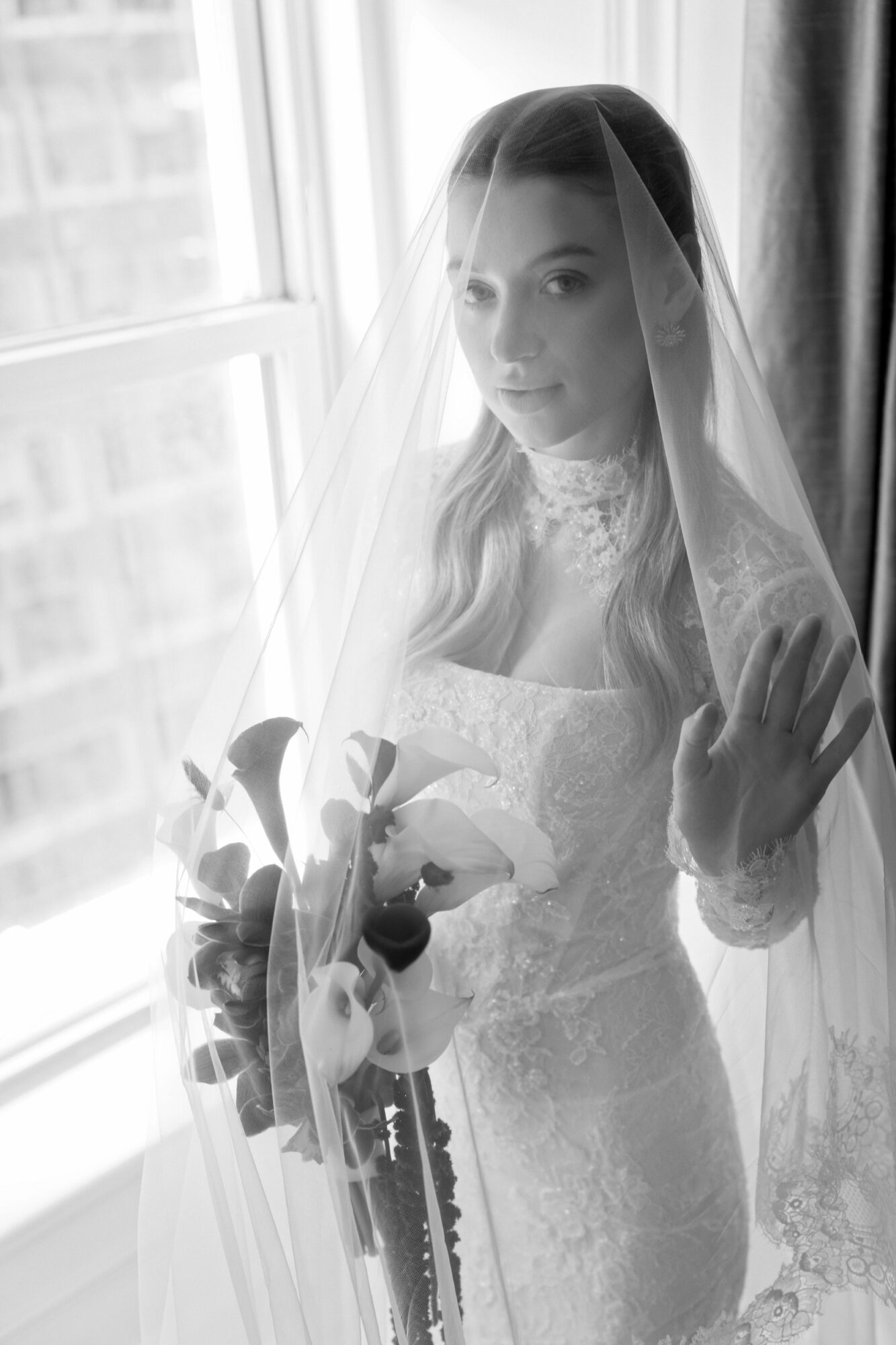 Bride with veil holding bouquet, standing by window, looking at camera, in a lace dress, black and white photo.