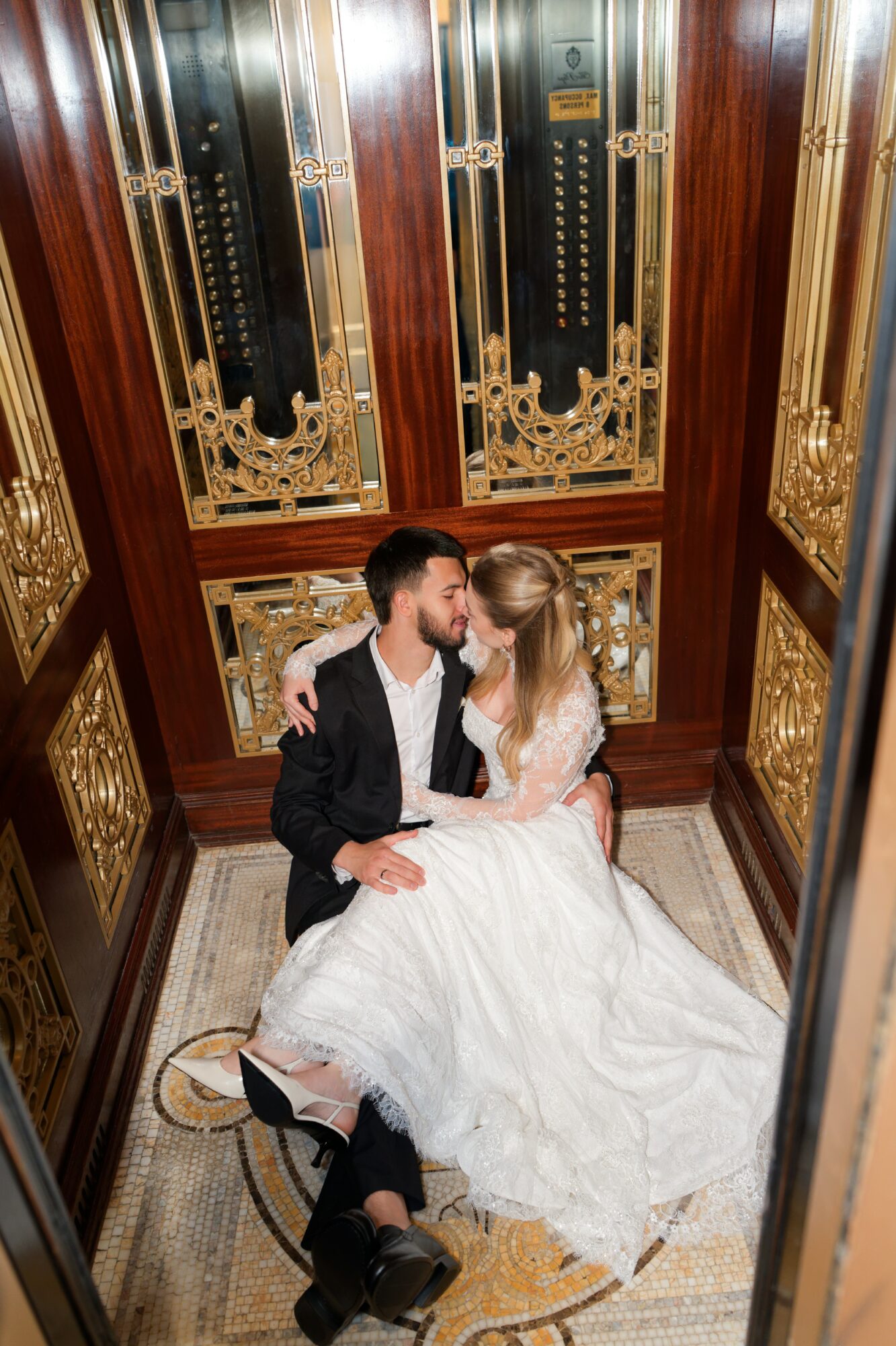 Couple sitting in elevator, woman in white dress, man in dark suit, facing each other, elevator decorated with gold and wood details.