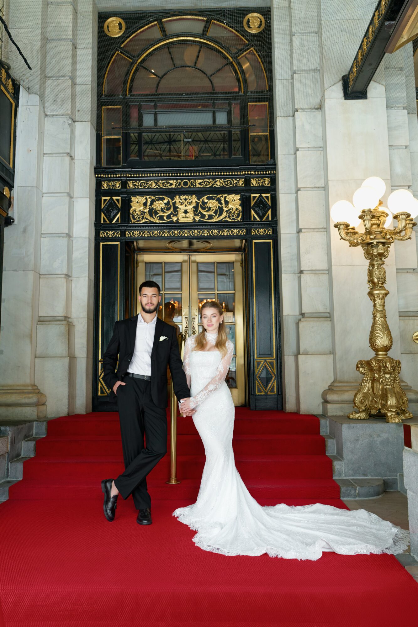 A couple in formal attire standing on a red carpet in front of a grand building entrance with ornate gold details and a large window.