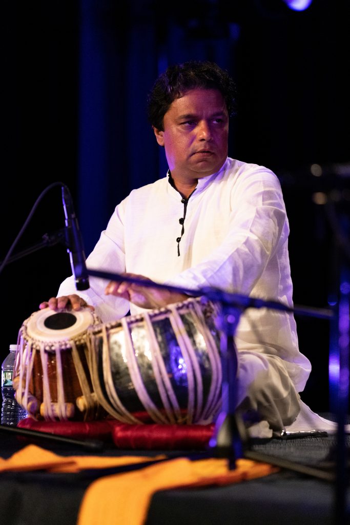 Man in white traditional attire playing tabla on stage with dark background.