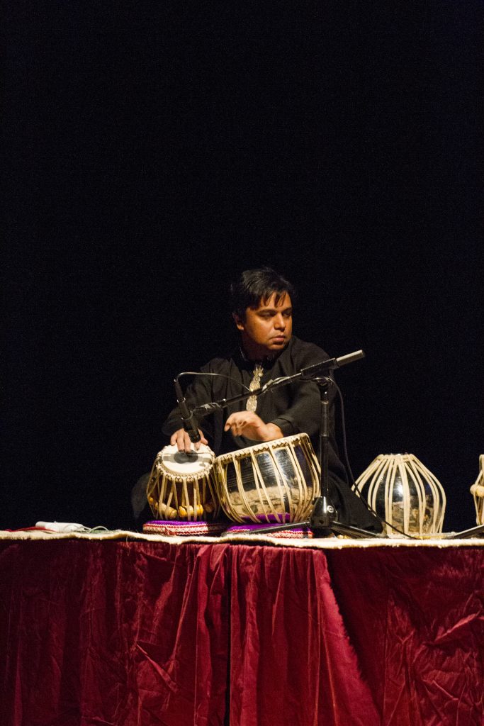 Person sitting behind a table with traditional drums, dark background, focused expression.