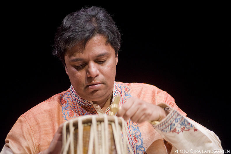 Person with short dark hair wearing an orange garment, focused on a traditional musical instrument against a black background.