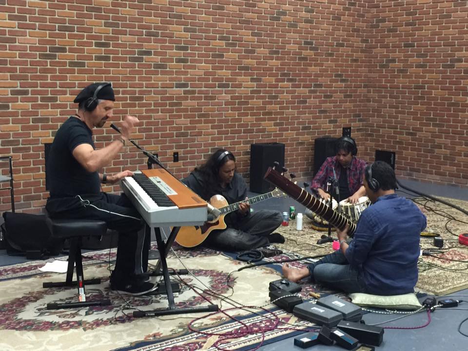 Four musicians perform with keyboard, guitar, and drums against a brick wall, seated on a patterned rug.