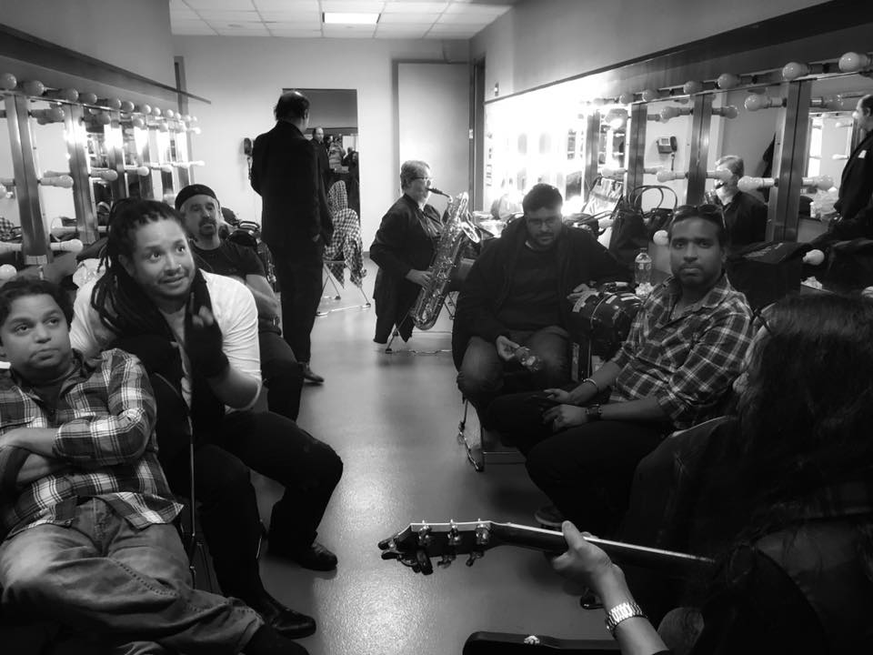 Group of people sitting and standing in a room with display cases, some holding musical instruments, in black and white.