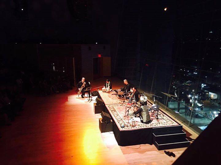 Musicians perform on a stage with a city view through large windows, illuminated by warm stage lighting.