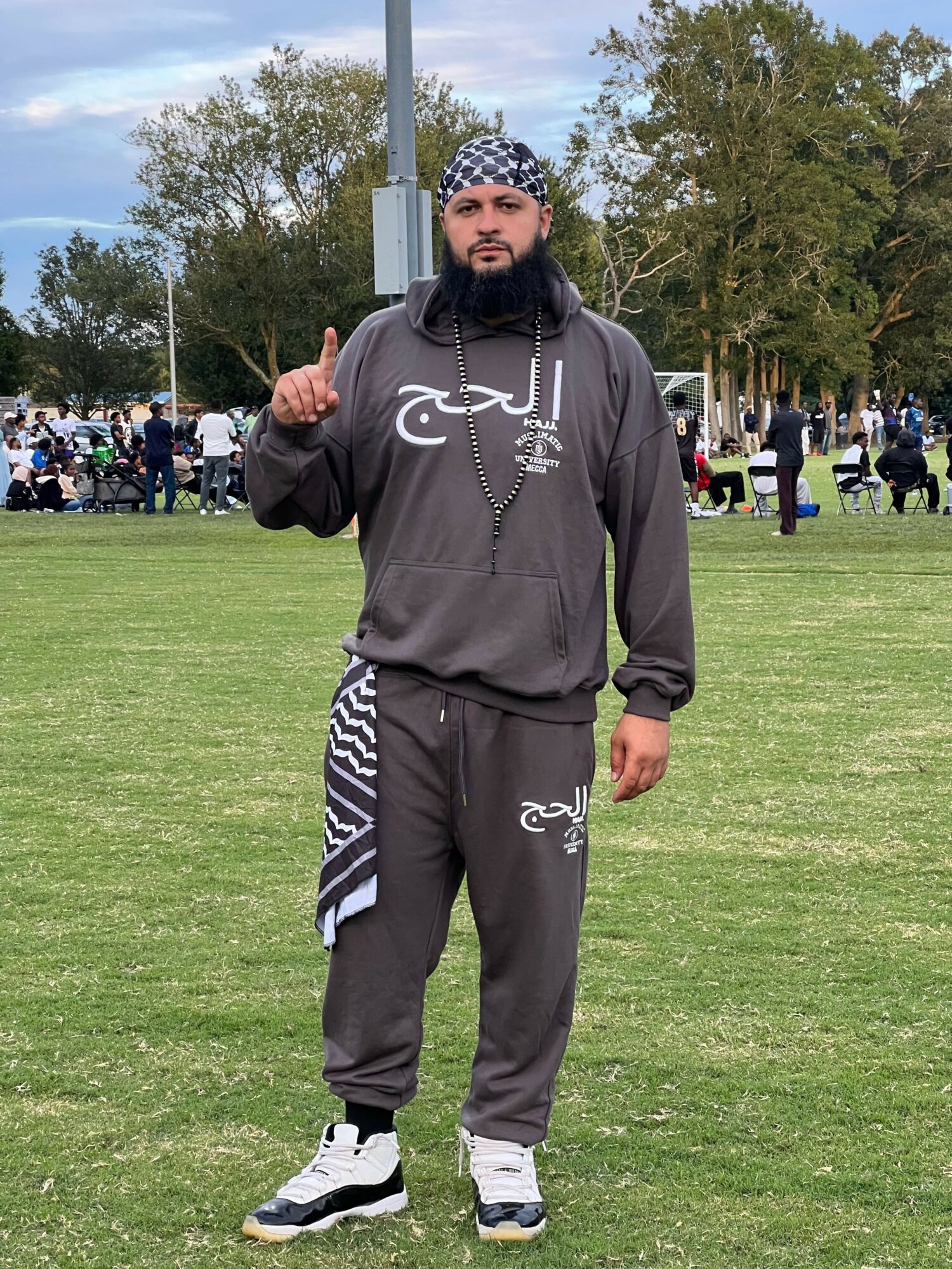 Person standing outdoors on grass, wearing a dark hoodie and pants, with trees and a crowd in the background.