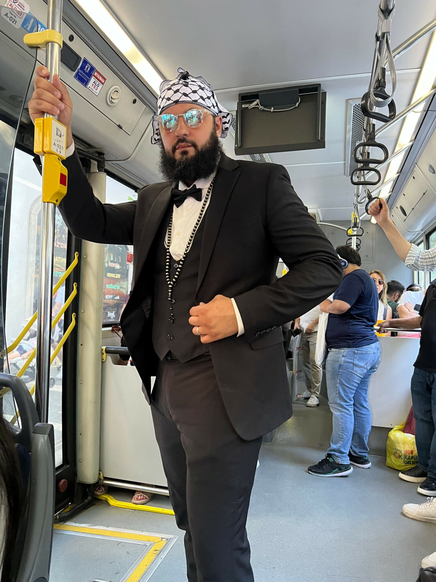 Man in black suit and face mask standing inside a bus, holding a yellow device, with other passengers in the background.