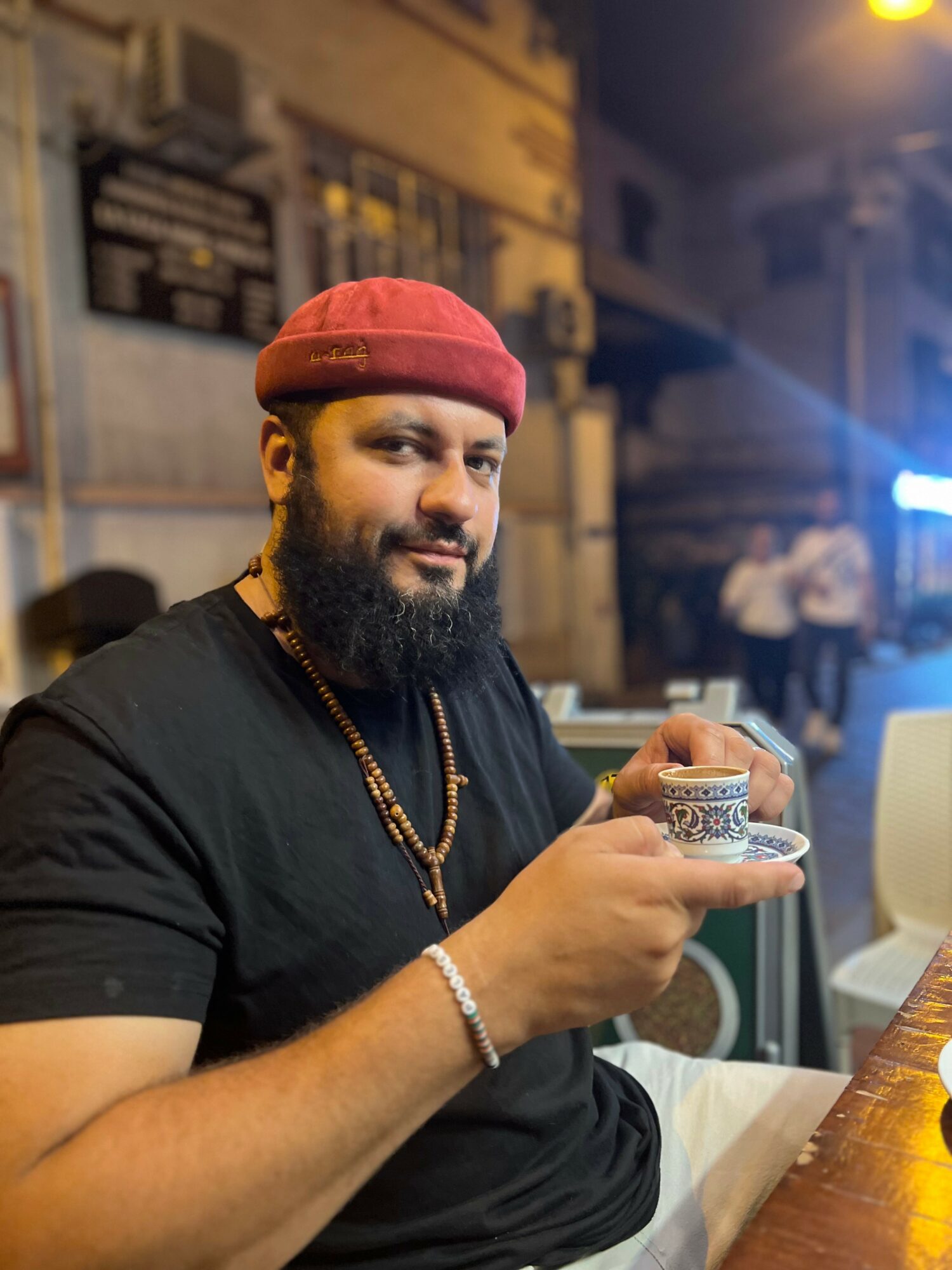 Man with a beard and red cap holding a glass, sitting at a table outdoors at night.