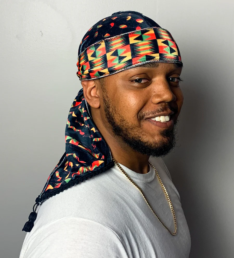 Smiling man with a colorful patterned durag and white shirt, standing against a plain wall.