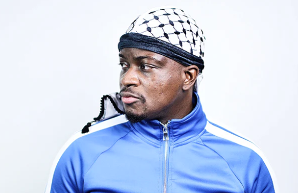 Man wearing a blue sports jacket and a patterned headscarf, looking to the side against a plain background.