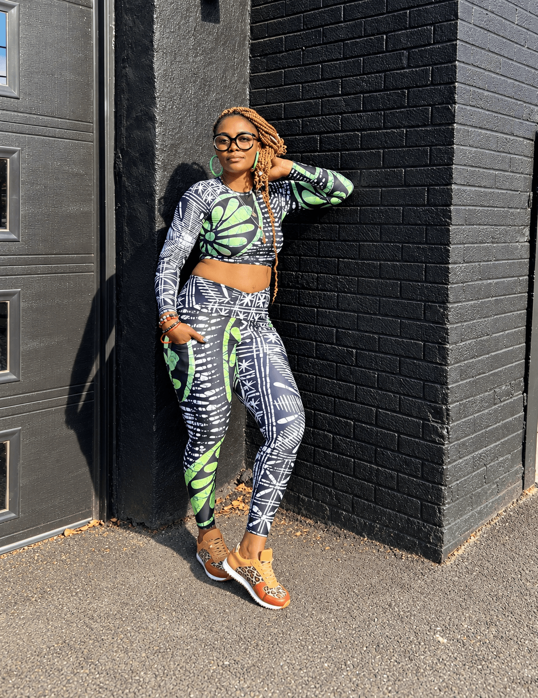 Woman with glasses and long hair standing against black brick wall, wearing colorful patterned outfit and orange sneakers.
