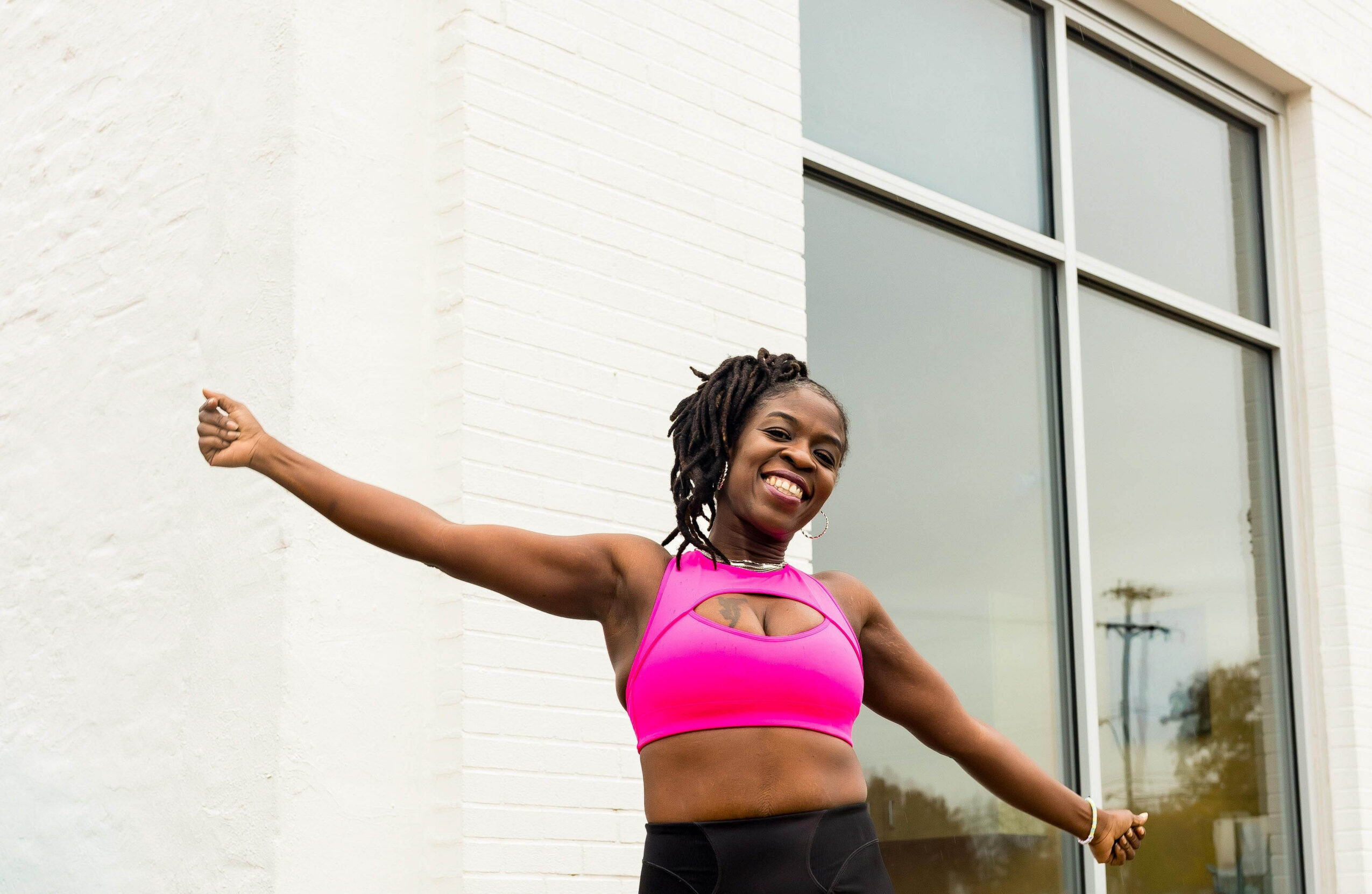 Woman with arms outstretched smiling outdoors near a building with large window, wearing a pink sports top.