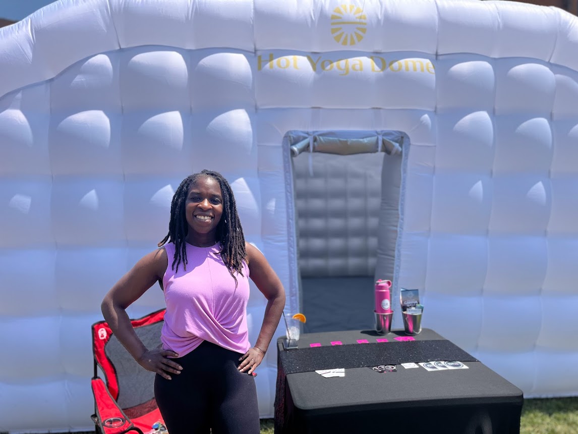 Woman in pink top and black pants standing in front of an inflatable structure, smiling with hands on hips.