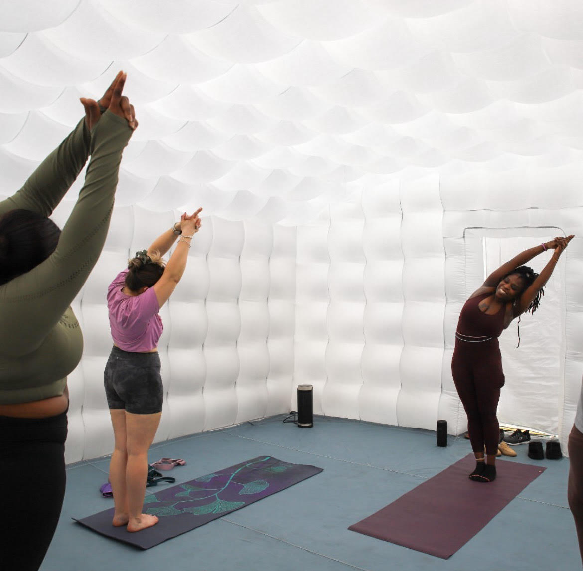 People practicing yoga in a white, textured room, standing on mats with arms raised and stretching.