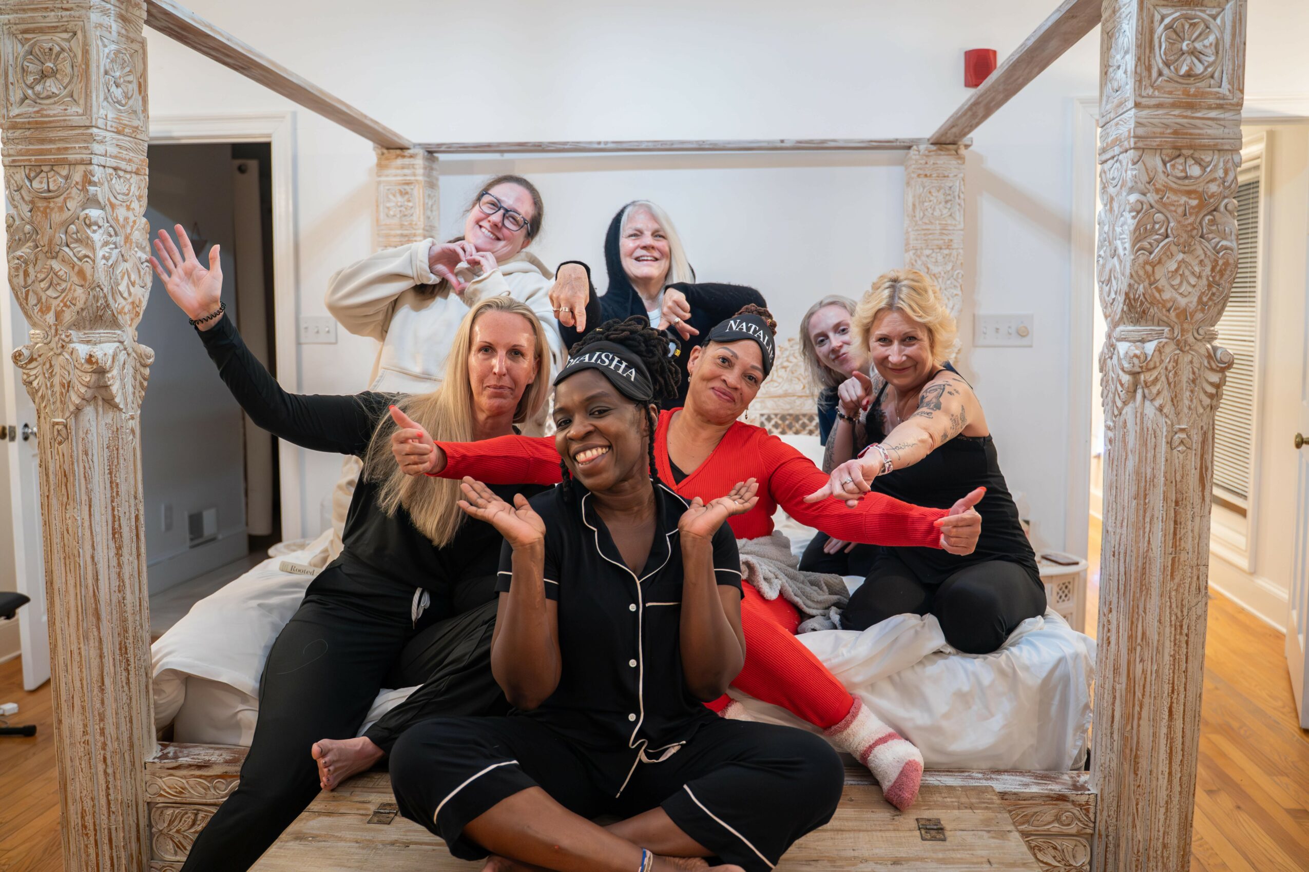 Group of six women sitting and posing on a bed in a decorated room, smiling and making gestures.