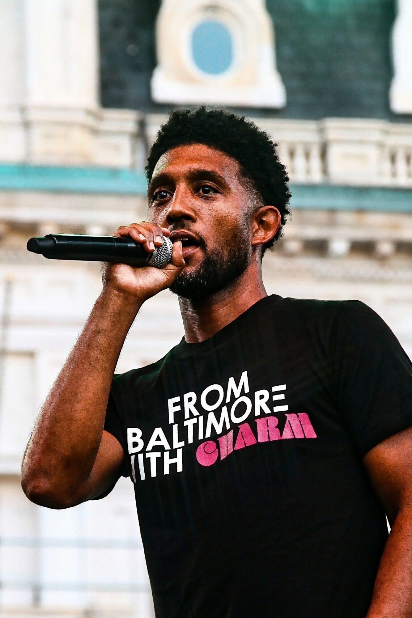 Man speaking into microphone outdoors, wearing a black T-shirt with text, background shows a building with architectural details.